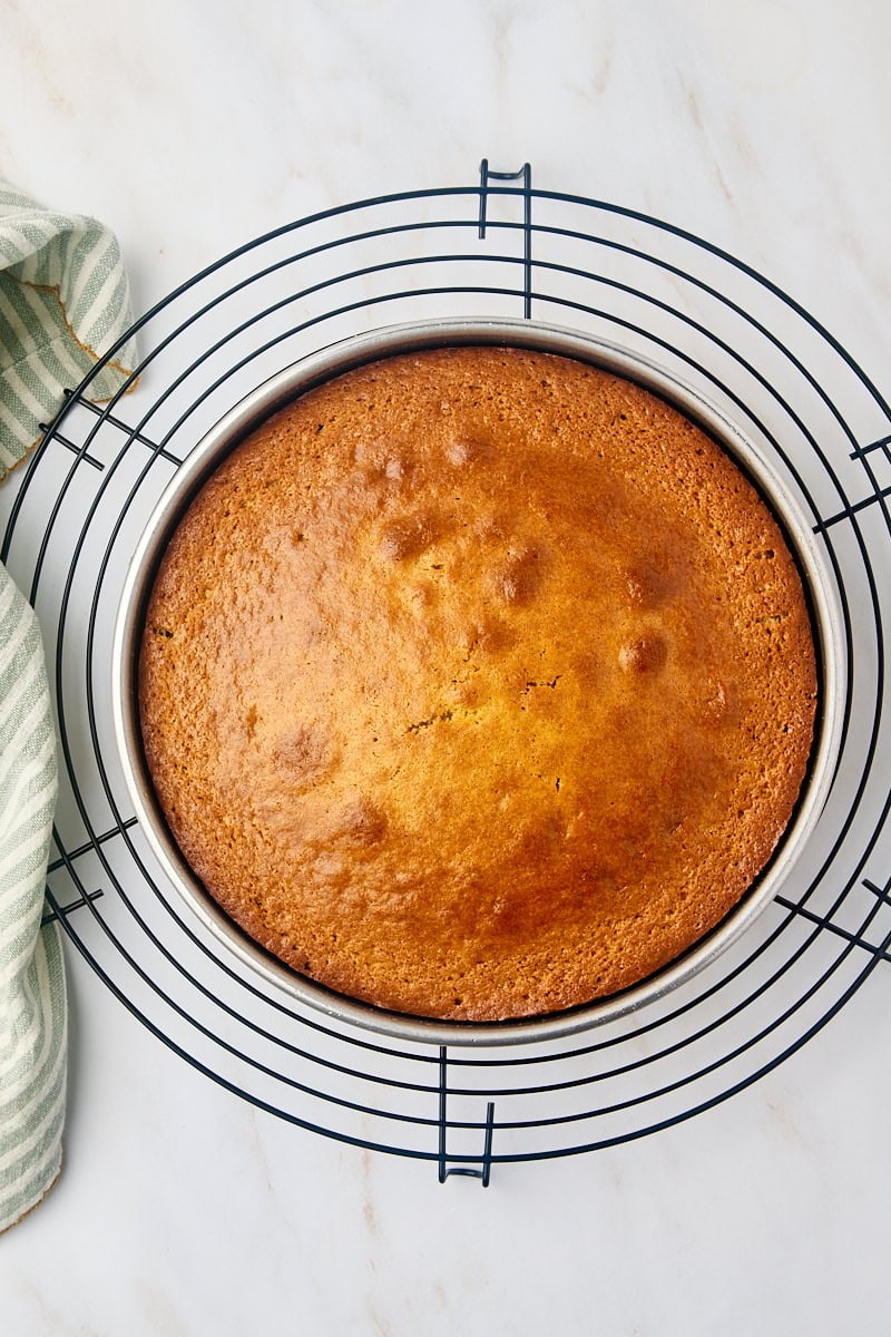 Overhead view of a freshly baked cardamom cake, still in its round pan, resting on a wire rack to begin cooling.
