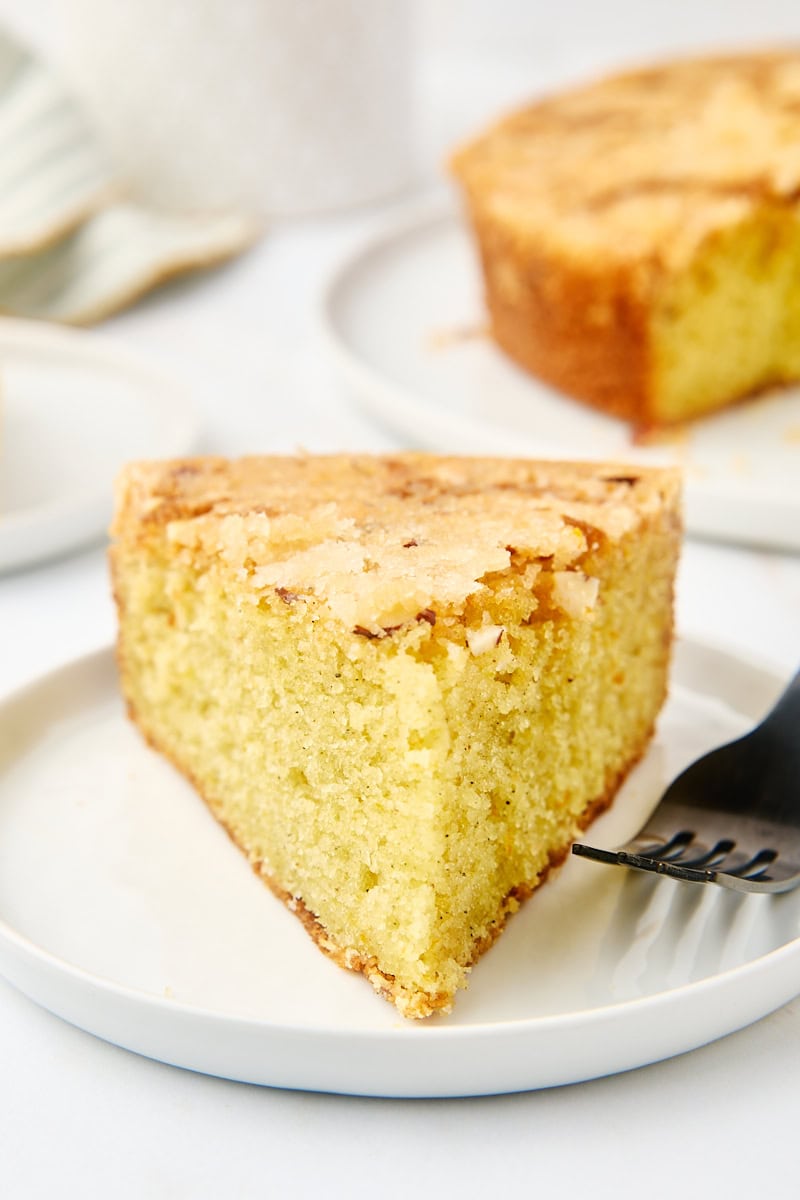 A slice of cardamom cake on a white plate with the remaining cake in the background, showing off the crunchy almond topping.