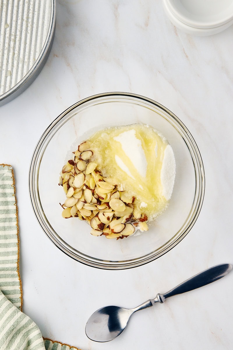 Overhead view of granulated sugar, sliced almonds, and melted butter being combined in a clear glass bowl for the cake topping.