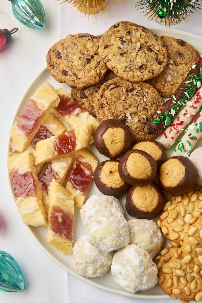 overhead view of various Christmas cookies and treats on a large white plate, surrounded by Christmas ornaments