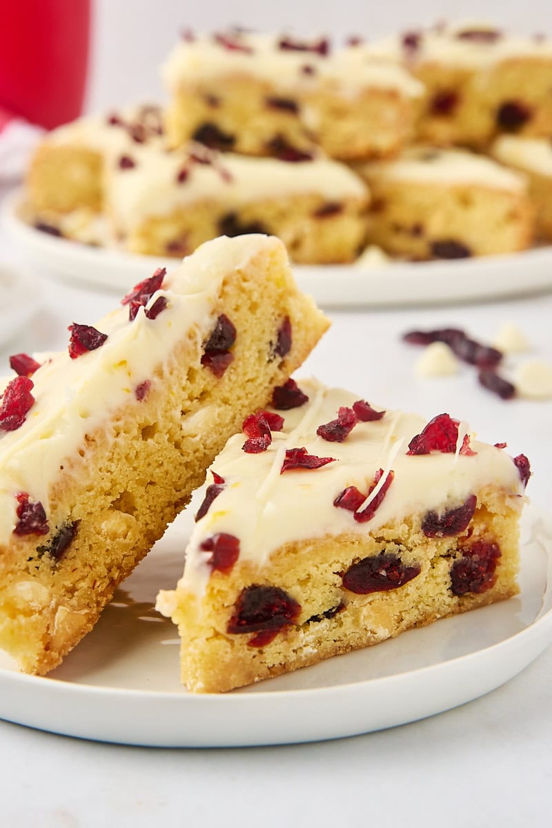 two cranberry bliss bars on a white plate with more bars on a large plate in the background