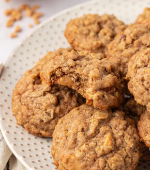 toffee almond oatmeal cookies piled on a white plate with a bite missing from the top cookie