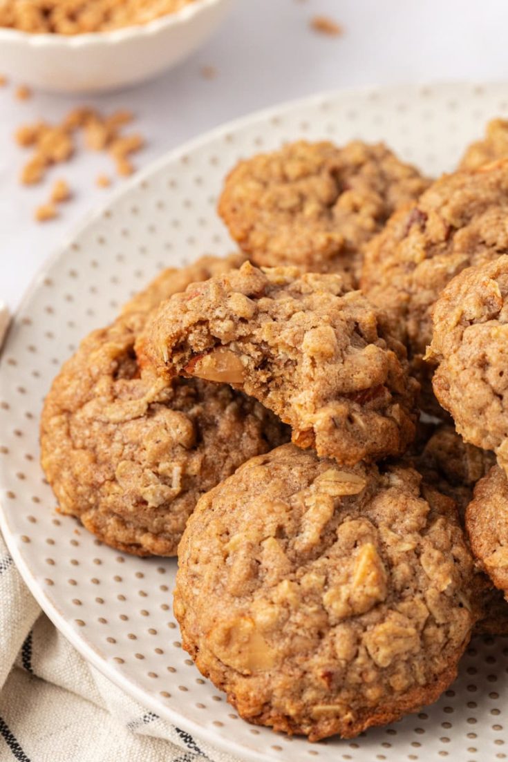 toffee almond oatmeal cookies piled on a white plate with a bite missing from the top cookie