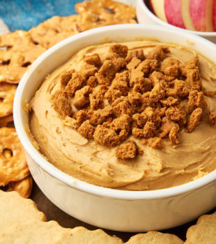 cookie butter dip in a white bowl surrounded by shortbread, pretzels, and apple slices