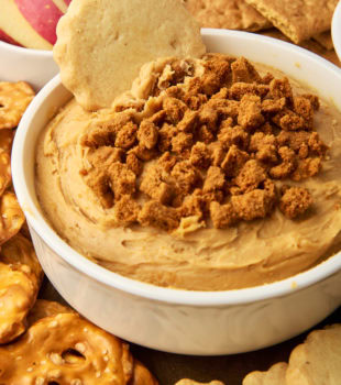 cookie butter dip in a white bowl surrounded by various foods for dipping