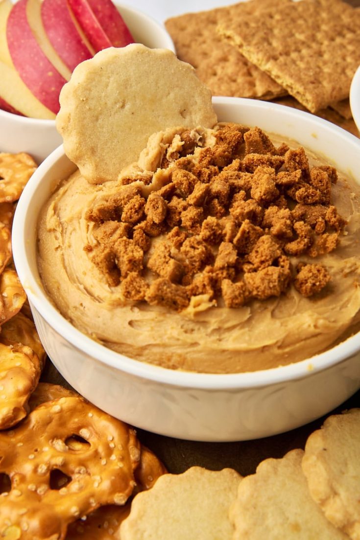 cookie butter dip in a white bowl surrounded by various foods for dipping