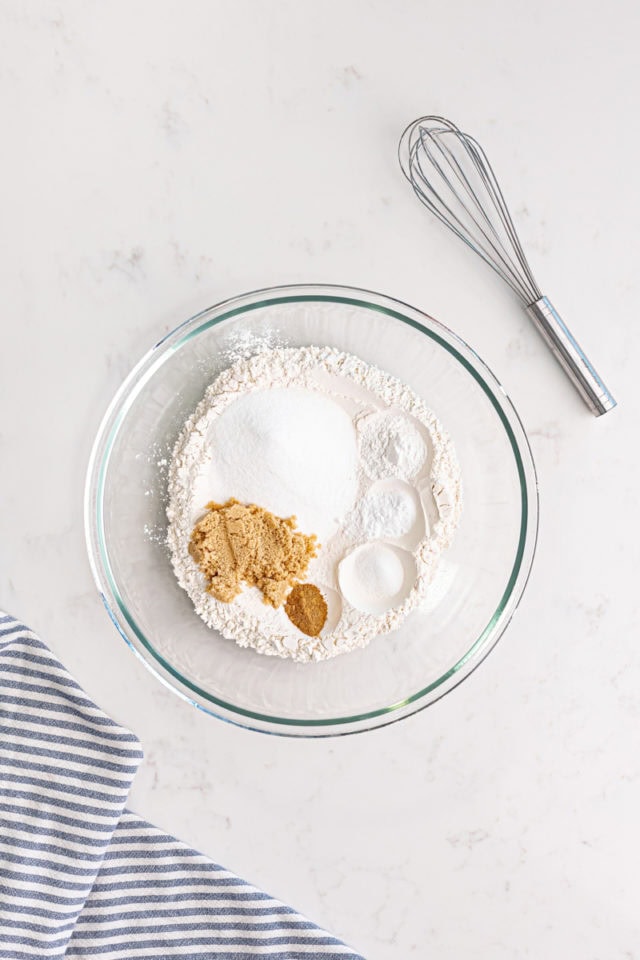 overhead view of flour, sugar, brown sugar, baking powder, baking soda, salt, and nutmeg in a glass mixing bowl