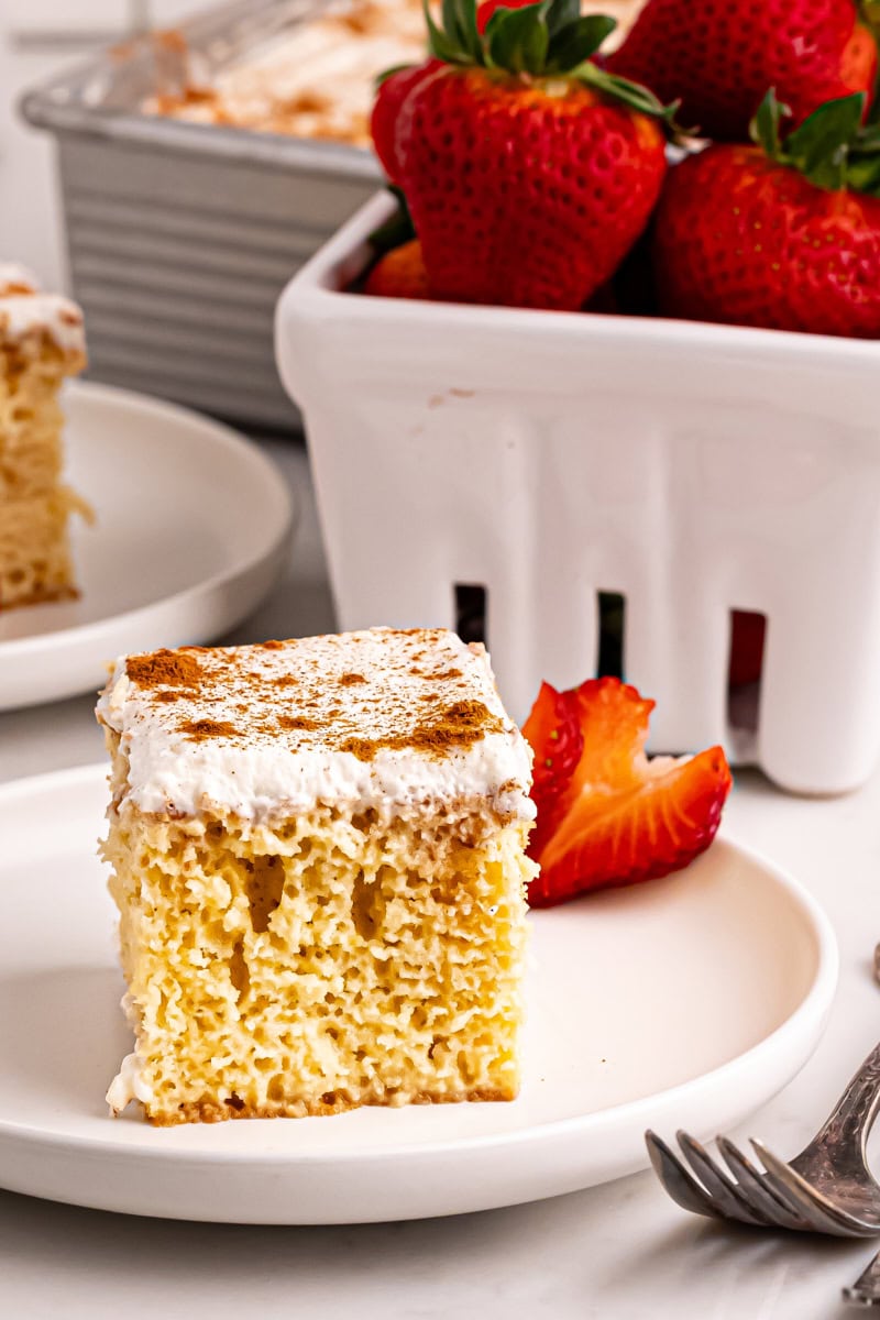 a slice of tres leches cake on a white plate with a basket of strawberries alongside