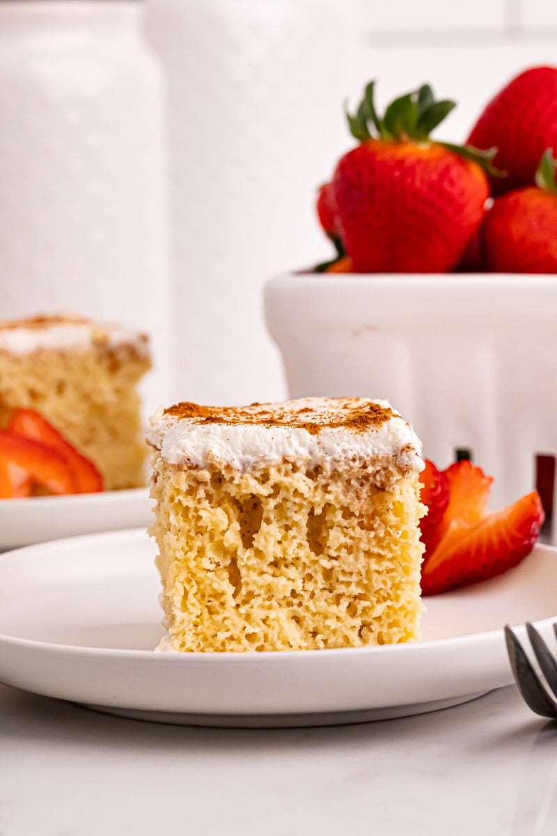 a slice of tres leches cake on a white plate with strawberries in the background