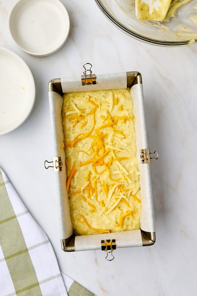 overhead view of shredded cheese sprinkled over the top of herb and cheese quick bread batter in a loaf pan