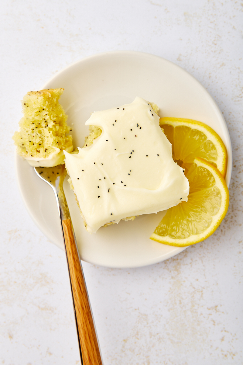 overhead view of a slice of lemon poppy seed cake with a bite on a fork, served on a white plate with two lemon slices