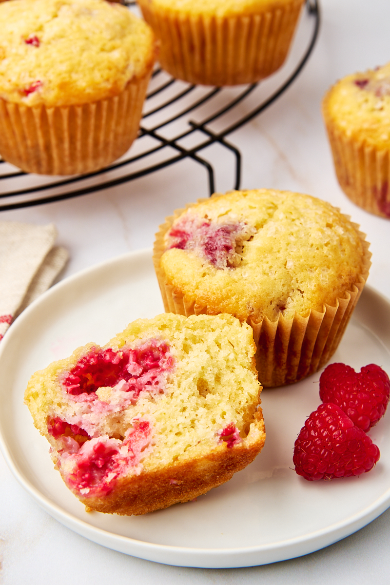 half of a lemon raspberry muffin and another muffin on a white plate with more muffins in the background