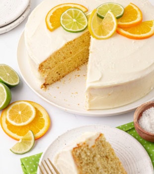 partially sliced citrus cake with tequila-lime frosting on a white cake plate alongside a slice on a white plate