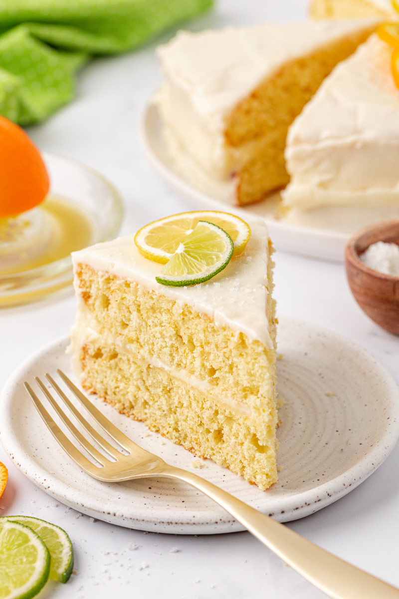 a slice of citrus cake with tequila lime frosting on a plate with remaining cake in the background