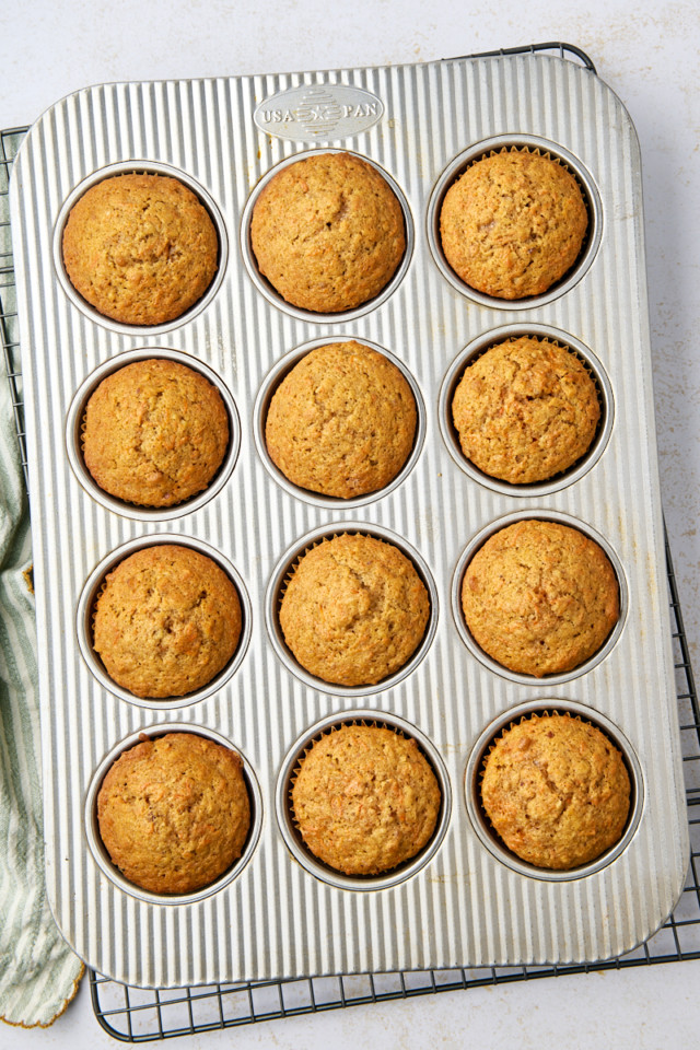 A metal muffin tin with twelve evenly baked carrot cake muffins sits on a cooling rack. Each muffin boasts a golden-brown top. A green cloth is partially visible on the side, set against a light countertop backdrop.