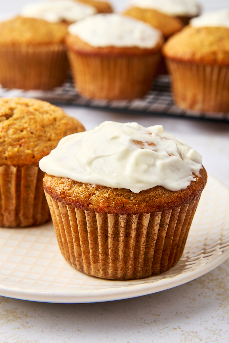 Close-up of a carrot cake muffin topped with a luscious cream cheese glaze on a white plate. Several unfrosted carrot cake muffins rest nearby on a cooling rack.