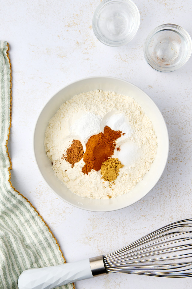 A white bowl containing flour, cinnamon, ginger, and baking powder on a light surface. A whisk with a white handle is nearby, alongside a green-striped cloth and two small glass bowls.