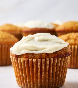 A close-up of a frosted cupcake with a luscious cream cheese glaze in the foreground, while a few unfrosted carrot cake muffins blur softly in the background. The frosting is creamy and white, contrasting with the golden brown cupcakes set on a light textured surface.