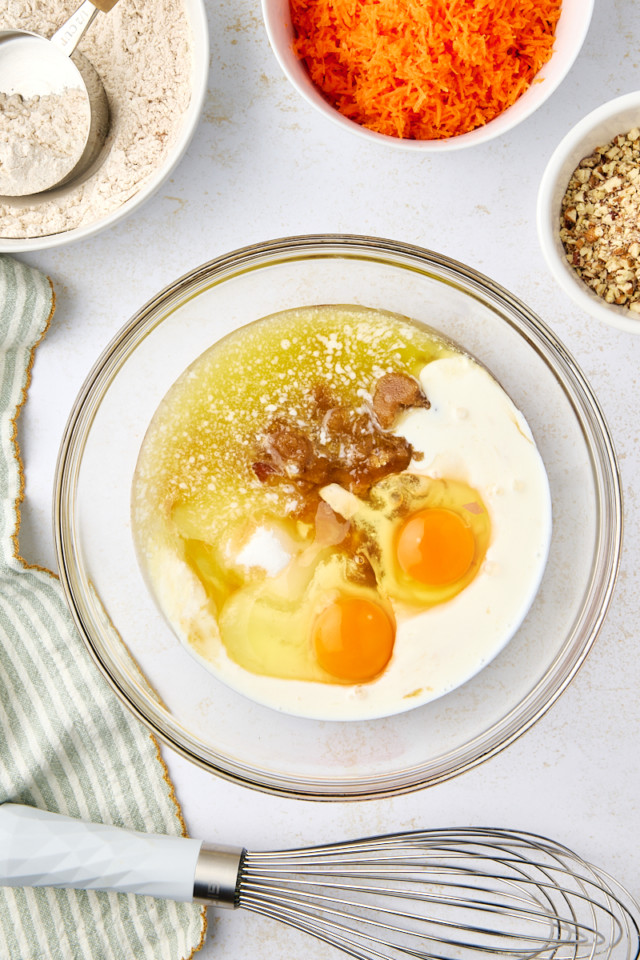 A glass bowl with eggs, milk, and other ingredients for a batter. Surrounding it are bowls of grated carrots, chopped nuts, flour, and a measuring cup. A whisk and a striped cloth are nearby on the white surface.