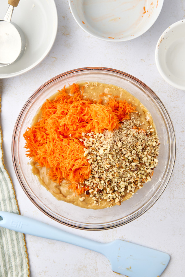 A glass bowl containing a mixture of shredded carrots, chopped nuts, and batter. Surrounding the bowl are several empty dishes, a measuring cup, and a blue spatula on a white surface. A striped cloth is partially visible on the left.