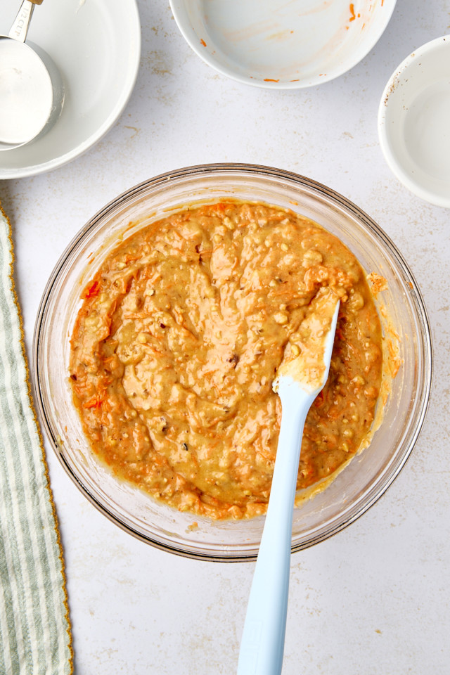 A glass bowl filled with thick, beige carrot cake muffin batter. A blue spatula rests inside the bowl. Nearby are empty white bowls, a measuring cup, and a striped cloth on a white surface.