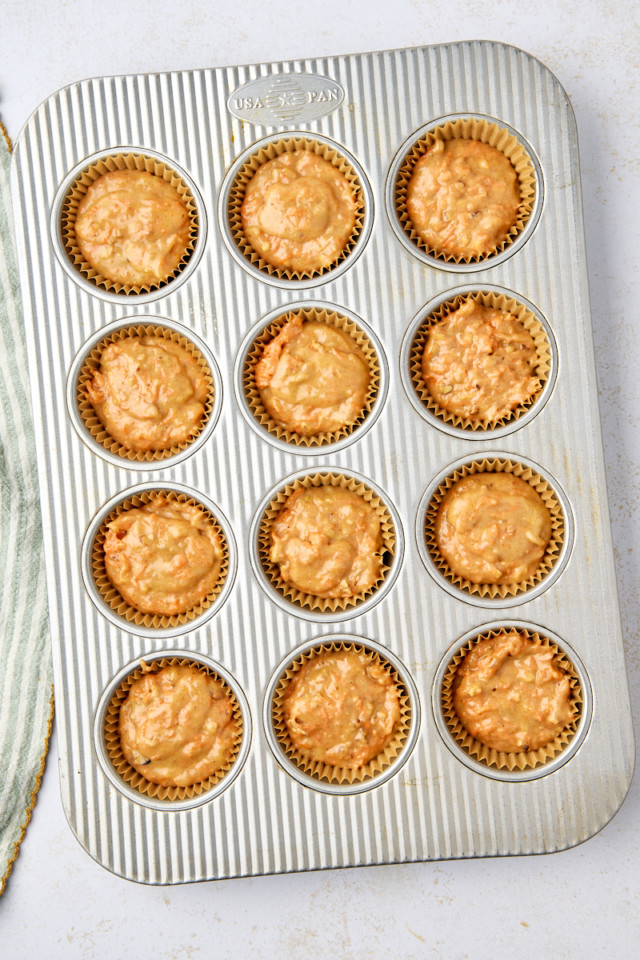 A muffin tray filled with 12 unbaked carrot cake muffins, each in a paper liner. The golden-brown muffins are evenly distributed and rest on a light-colored surface. A striped cloth is partially visible on the side, adding a touch of charm.