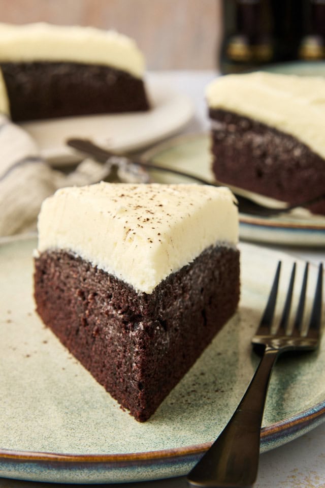 A slice of rich Guinness chocolate cake with creamy frosting rests on a green plate, accompanied by a fork. The cakes deep, dark texture contrasts beautifully with the light cream cheese frosting layered on top. More delectable slices await in the background.