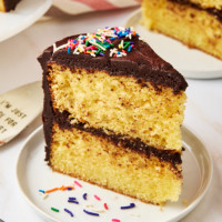 a slice of yellow cake with chocolate frosting on a white plate with sprinkles