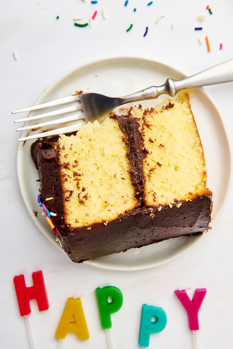 overhead view of a partial slice of yellow cake with chocolate frosting on a white plate with a fork; letter-shaped candles spelling out 