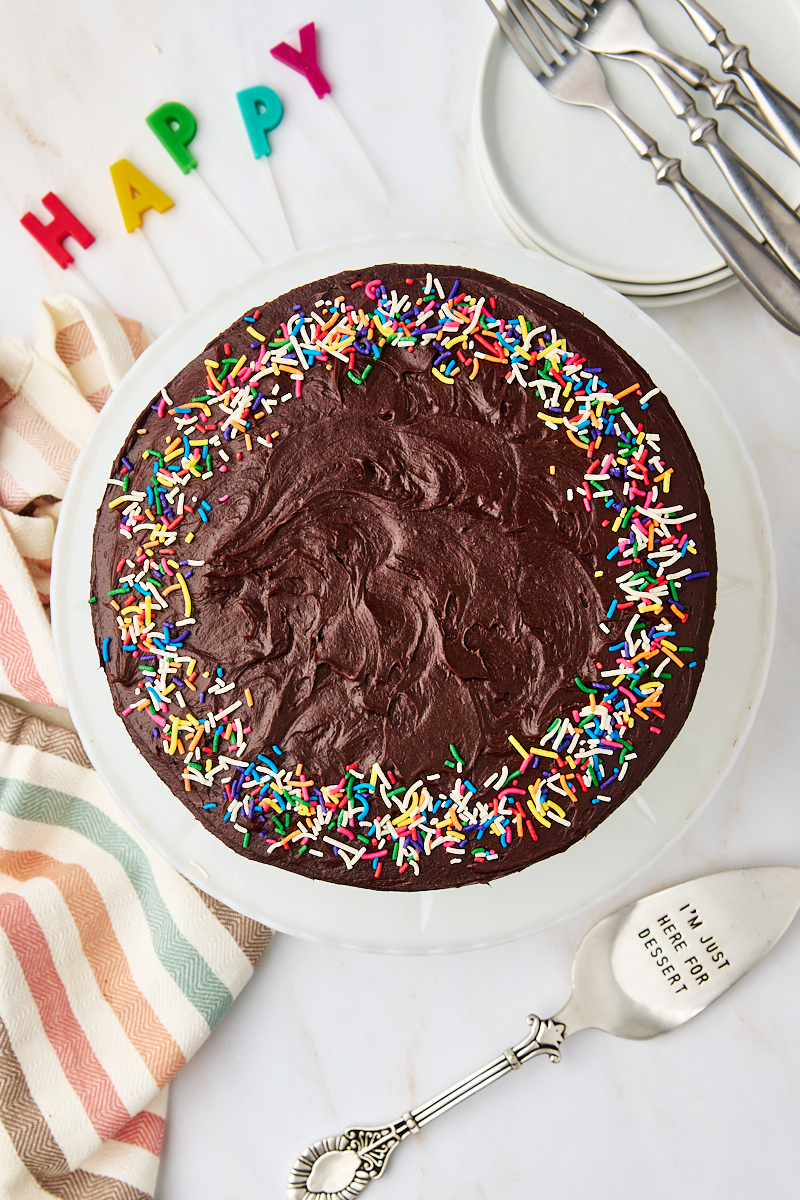 overhead view of yellow cake with chocolate frosting on a white cake plate, surrounded by plates, forks, a cake server, and birthday candles