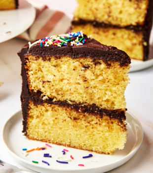 a slice of yellow cake with chocolate frosting on a white plate with more cake in the background