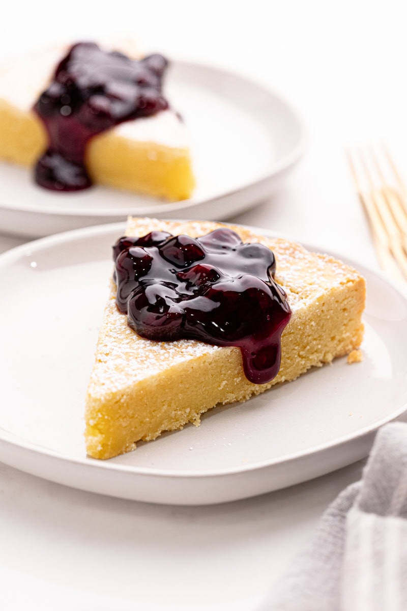 a slice of almond gÃ¢teau Breton topped with blueberry compote and served on a white plate; another served slice in the background