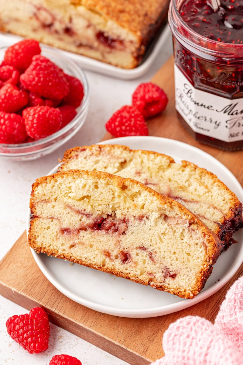 two slices of berry swirl pound cake on a white plate surrounded by fresh raspberries, more cake, and a jar of raspberry preserves
