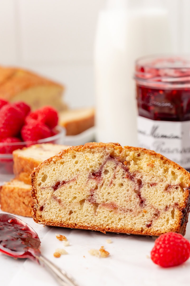 a slice of berry swirl pound cake with more cake, raspberries, and a jar of raspberry preserves in the background