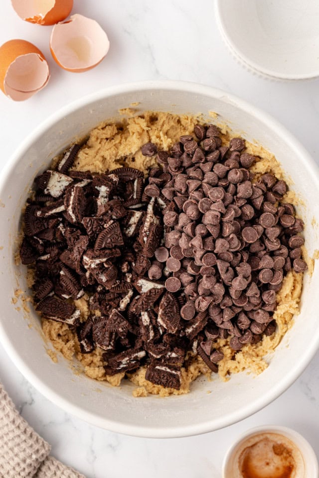overhead view of chocolate chips and chopped Oreos added to cookie dough in a mixing bowl