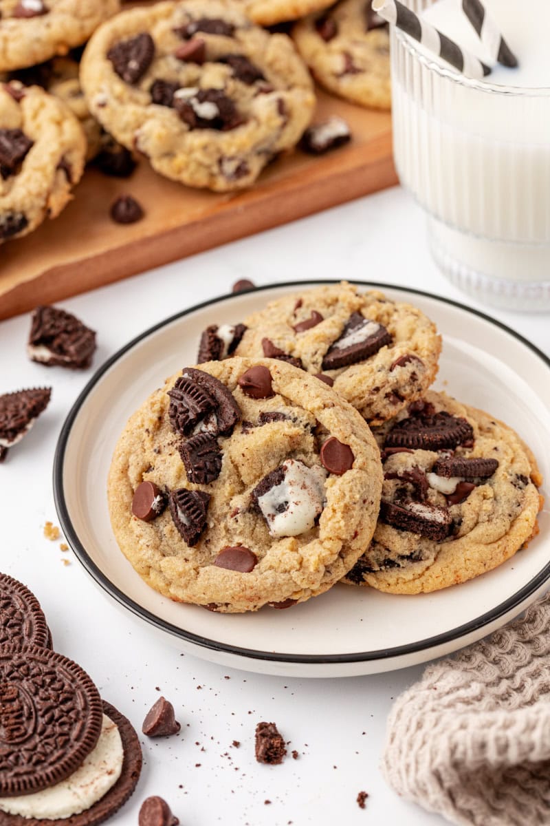three Oreo chocolate chip cookies on a white plate with more cookies and a glass of milk in the background