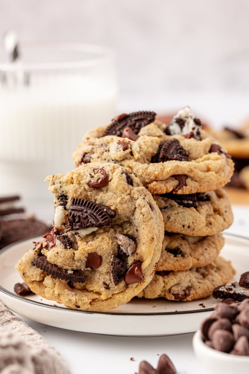 an Oreo chocolate chip cookie with a bite missing leaned against a stack of four more cookies; a glass of milk partially visible in the background