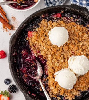 berry crisp in a skillet, with some already served and a spoon resting in the pan; a serving of crisp partially visible in the top left corner