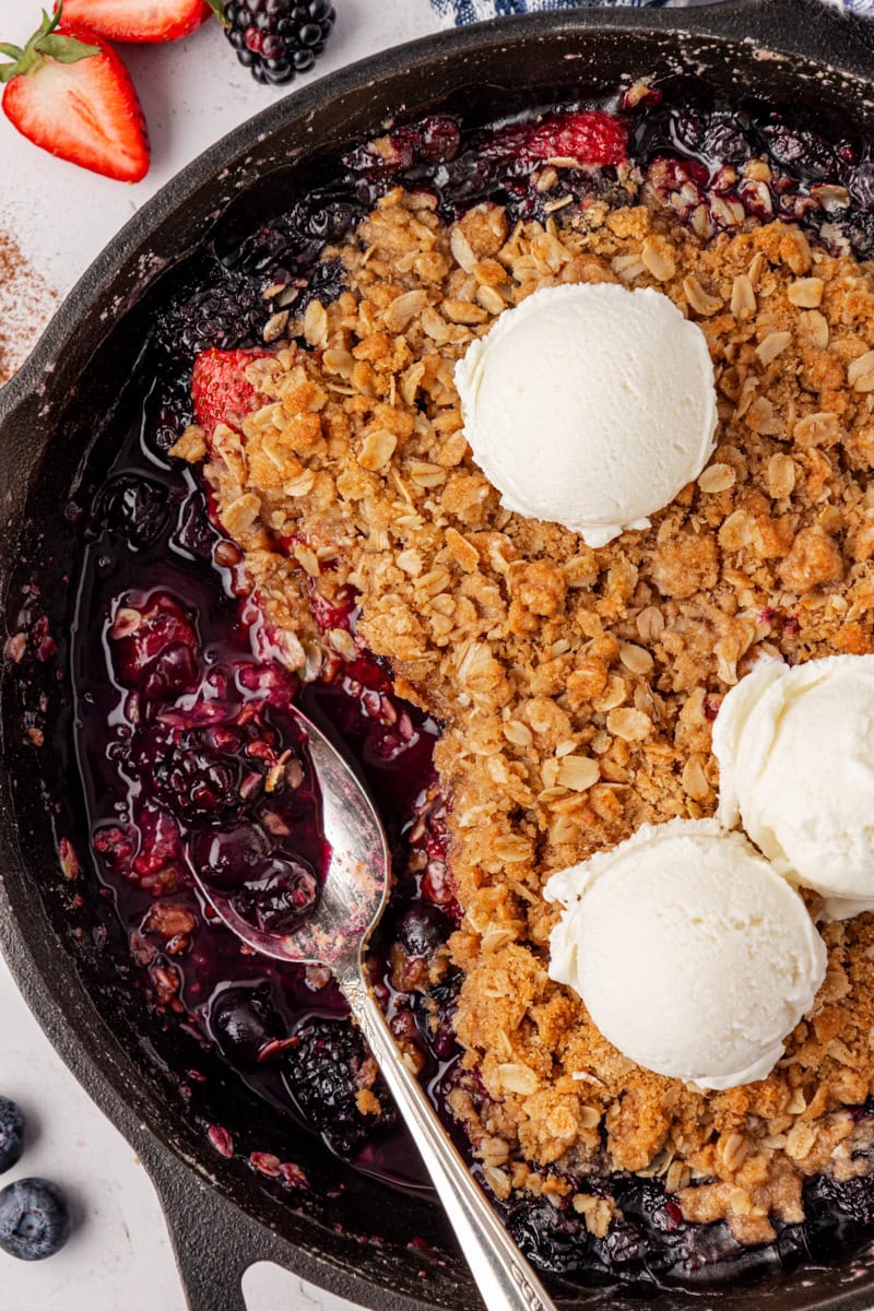 a spoon resting in a skillet where berry crisp has been scooped out; remaining crisp is topped with three scoops of ice cream