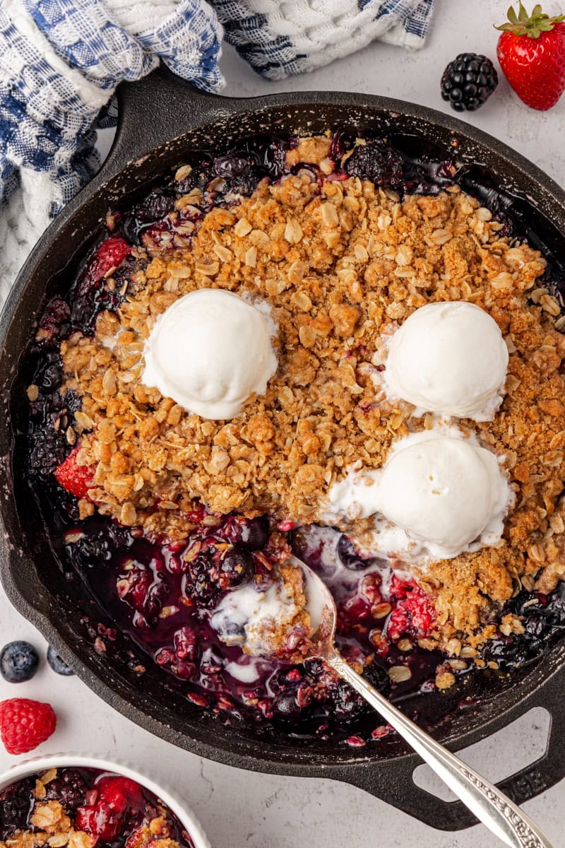 overhead view of berry crisp in a cast iron skillet, topped with three scoops of ice cream; a spoon rests inside the skillet where some of the crisp has been removed