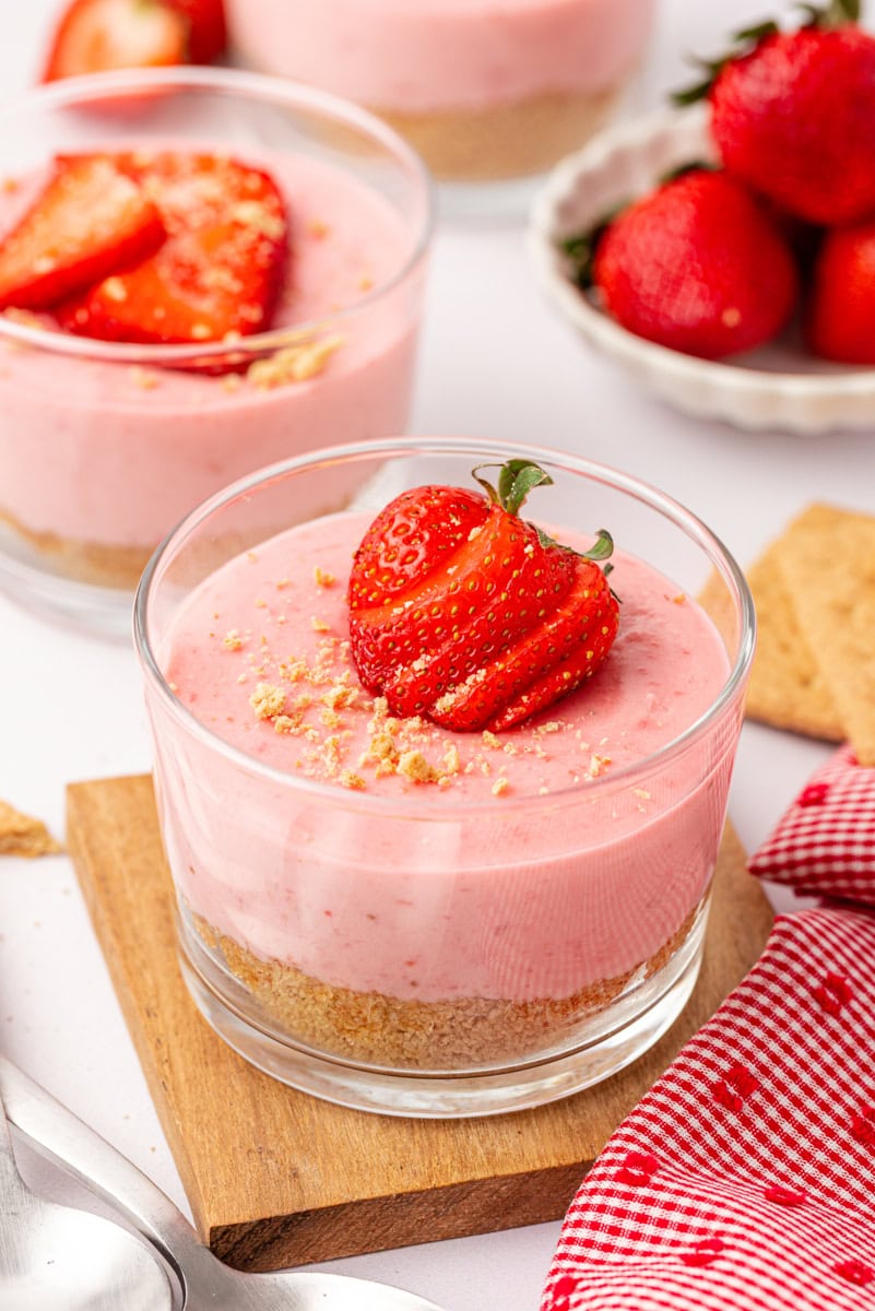 a strawberry no-bake cheesecake in a clear glass with more cheesecakes and a bowl of strawberries partially visible in the background