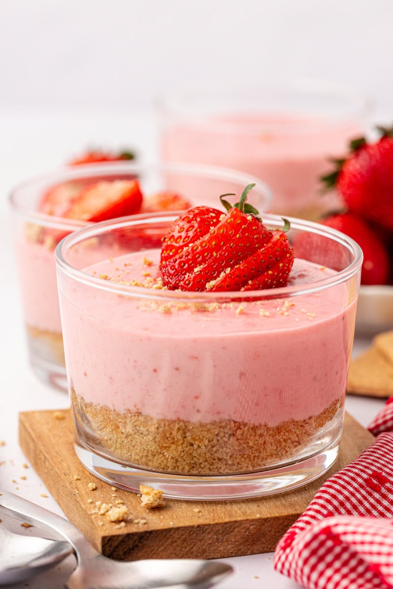 a strawberry no-bake cheesecake in a clear glass on a wooden plate; more cheesecakes in the background