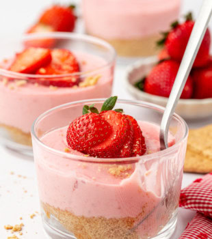 a strawberry no-bake cheesecake with a spoon sticking out of its jar; more cheesecakes and a few strawberries partially visible in the background