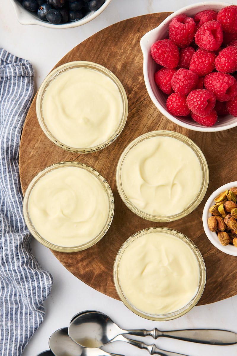 overhead view of four servings of white chocolate mousse in glass ramekins on a wooden board surrounded by fresh berries, spoons, and a blue striped napkin