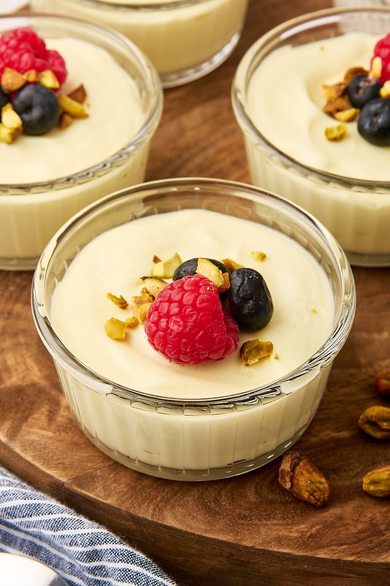 a serving of white chocolate mousse topped with raspberries, blueberries, and pistachios served in a clear ramekin; more servings partially visible around it