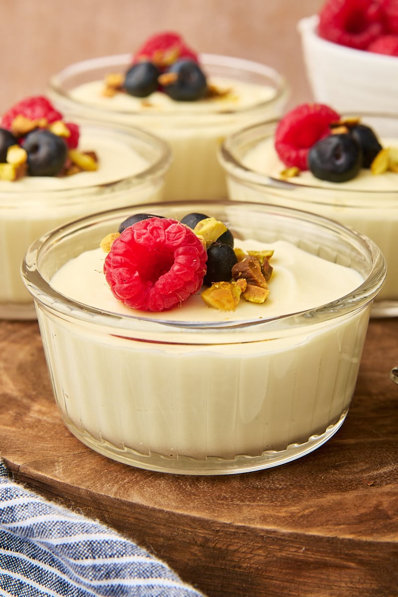 Four servings of white chocolate mousse in clear ramekins set on a wooden board. Each is topped with fresh berries and chopped pistachios.
