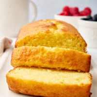 a partially sliced yogurt cake on a white serving plate; fresh berries and a coffee mug partially visible in the background