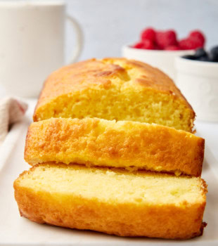 a partially sliced yogurt cake on a white serving plate; fresh berries and a coffee mug partially visible in the background