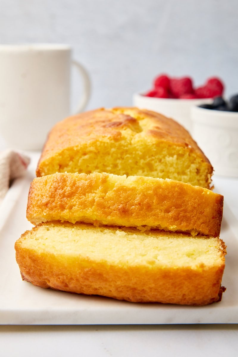 a partially sliced yogurt cake on a white serving plate; fresh berries and a coffee mug partially visible in the background