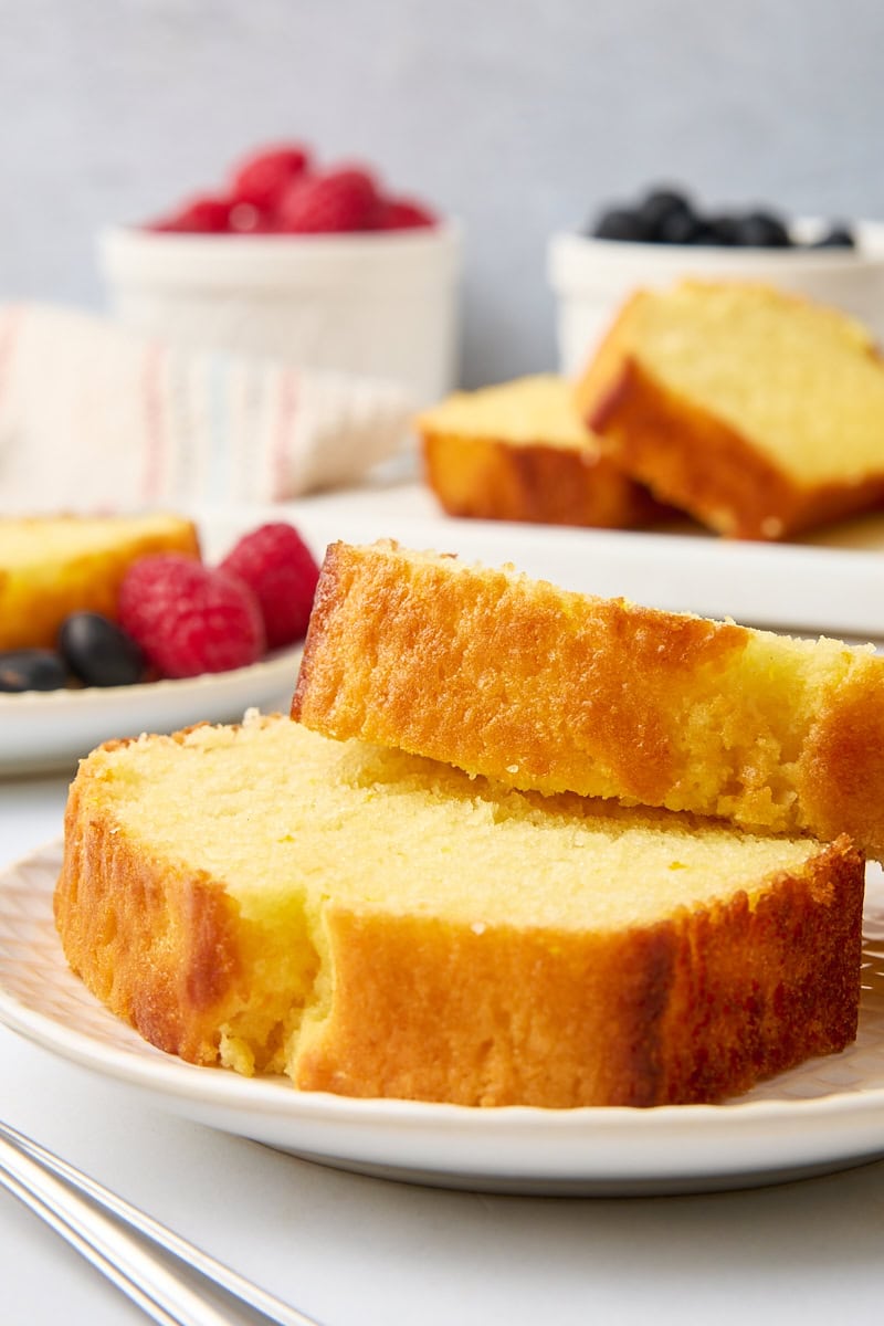 two slices of yogurt cake on a plate with more cake and bowls of fresh berries in the background